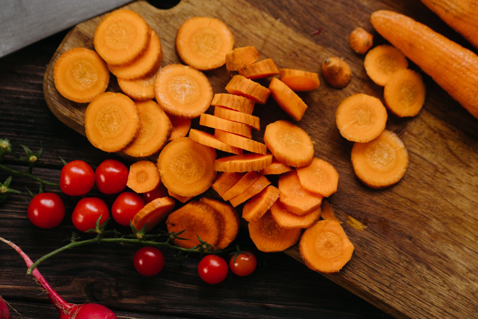 Rodelas e pedaços de cenoura sobre tábua de madeira, com tomates ao lado.